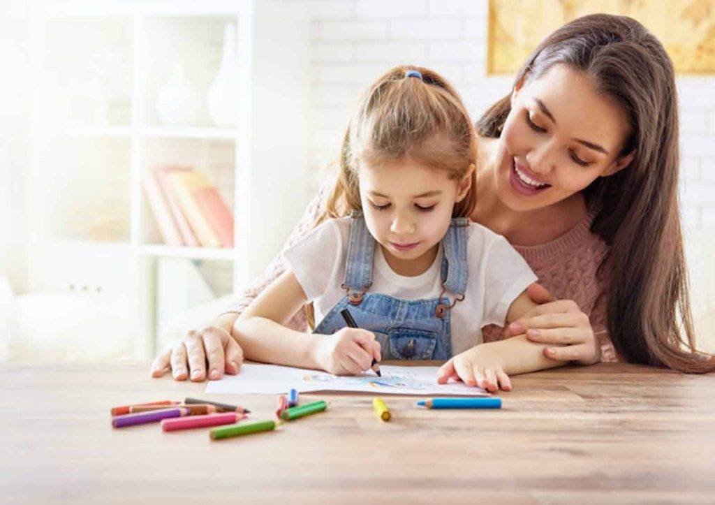 A woman, possibly a BCBA Miami therapist, smiles and hugs a young girl as the girl colors with colored pencils at a wooden table. The bright, cheerful scene is filled with art supplies spread out on the table.
