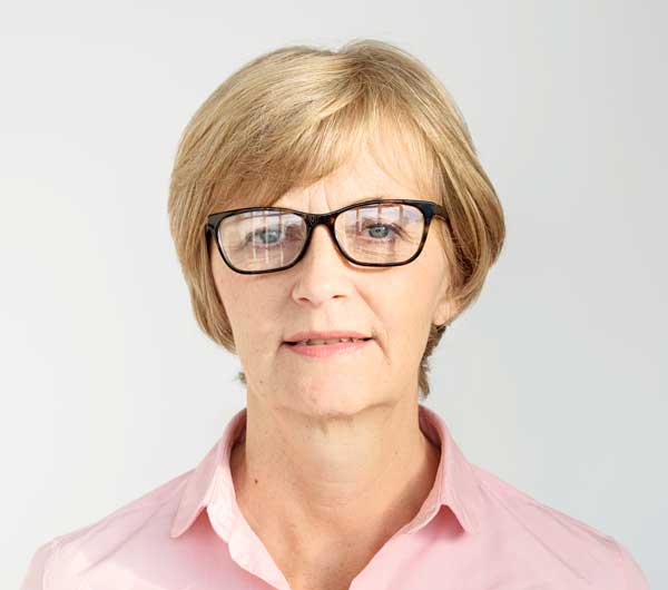 A middle-aged woman with short blonde hair and glasses, wearing a light pink collared shirt, stands against a plain light background. She looks directly at the camera with a neutral expression, embodying the professionalism of a BCBA Miami specialist.