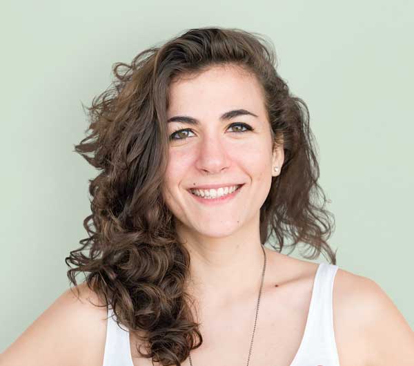 A young woman with wavy brown hair and light skin smiles at the camera. She is wearing a white tank top and a necklace, standing against a plain light green background, embodying the confident spirit of a BCBA Miami professional.
