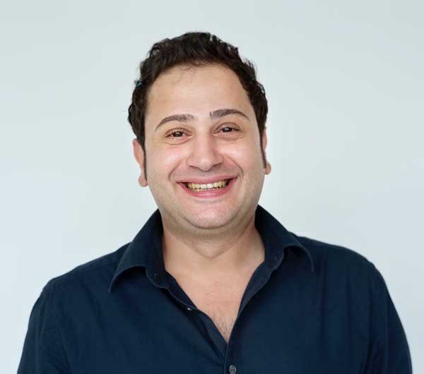 A man with dark hair smiles warmly at the camera. He is wearing a dark blue shirt over a white background.